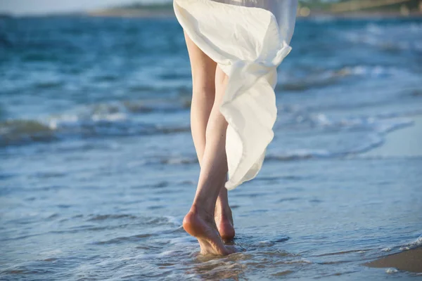 一个穿着白色连衣裙的漂亮女人在日落时沿着海滩边散步 地平线线 图库图片