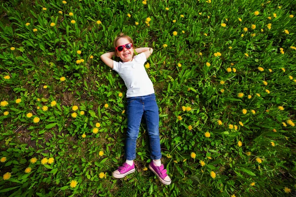 Liten flicka ligger på en grön gräsmatta med gula maskrosor, ovanifrån — Stockfoto