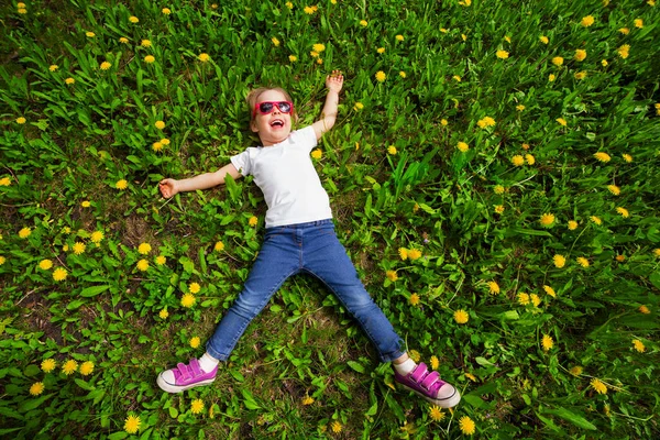 Klein meisje ligt op een groen gazon met gele paardebloemen, bovenaanzicht — Stockfoto