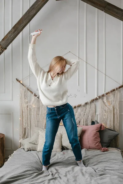 Glad ung kvinna i hörlurar lyssnar på musik från smartphone och dansar på sängen hemma. — Stockfoto