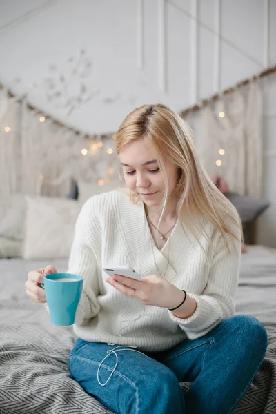 Joven mujer caucásica se sienta en la cama y utiliza un teléfono inteligente . — Foto de Stock