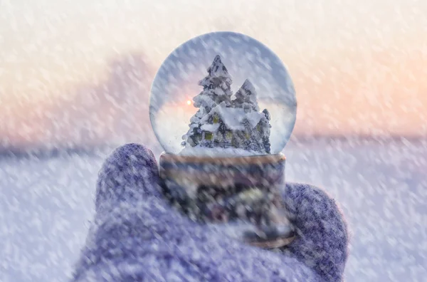 Fille en mitaines tricotées tenant boule de verre avec sapins, maison et neige artificielle avec paysage d'hiver avec chute de neige à l'arrière-plan — Photo