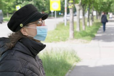 Siyah şapkalı ve sağlık maskesi takan çekici bir kadın güneşli bir bahar gününde şehir caddesinde yürüyor.
