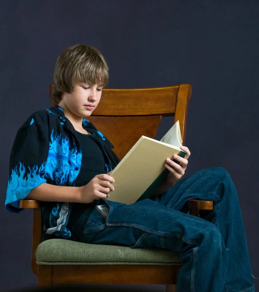 Young boy reading while seated in a wooden chair. — Stock Photo, Image