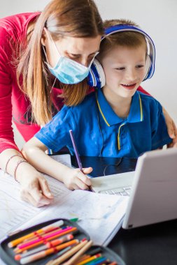 Coronavirus salgını. Kilitlenme ve okul kapanması. Anne, oğlunun yüz maskesi takmasına evde online ders çalışmasına yardım ediyor. COVID-19 salgını çocukları ve öğretmenleri çevrimiçi öğrenmeye zorluyor.