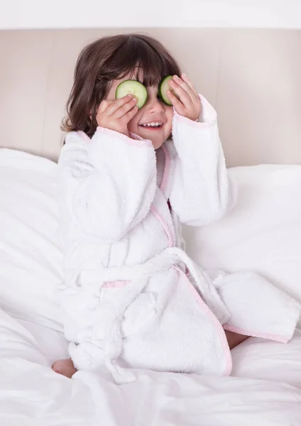 Hübsches Mädchen im Bademantel mit Gurkenscheiben auf dem Bett — Stockfoto