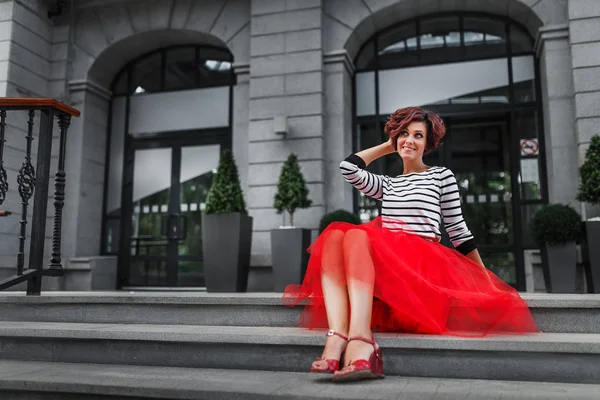 Young charming dark-haired woman in bright red skirt sits on the steps of the city staircase — Stock Photo, Image