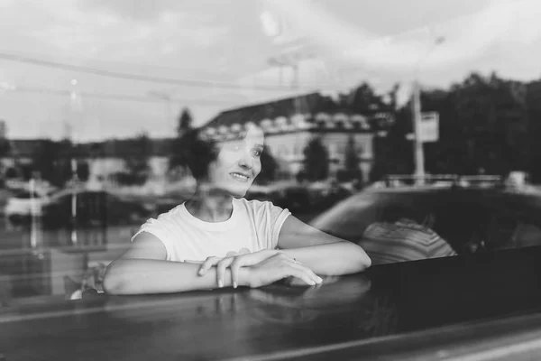 Atractiva joven sentada a la mesa en la cafetería. Fotografiado desde la calle con reflejos de la ciudad en la ventana —  Fotos de Stock