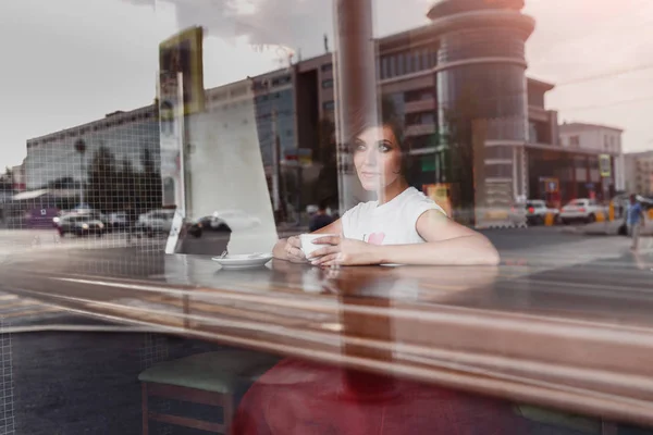 Όμορφη κοπέλα απολαμβάνοντας ένα ζεστό καφέ ή τσάι μέσα σε ένα καφέ στην πόλη. Πυροβόλησε μέσα από το γυαλί με αντανακλάσεις — Φωτογραφία Αρχείου