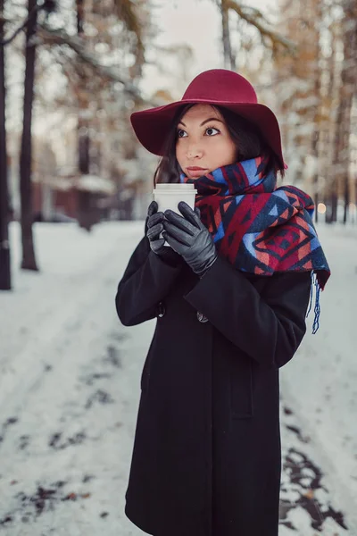 Outdoor portrait of young Caucasian woman, holding a cup of takeaway coffee in winter park on snowy day. Dressed in an elegant wide-brimmed hat and bright scarf — Stock Photo, Image