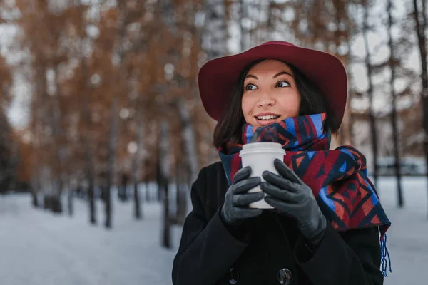 Woman with big mug of hot takeaway drink during cold day. Dressed in wide-brimmed hat and bright scarf