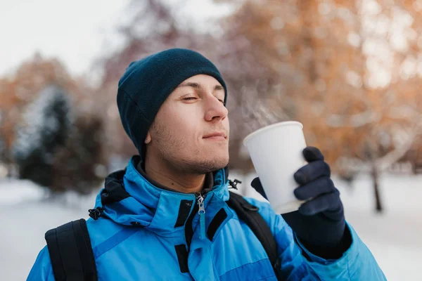 Close-up de um homem de chapéu quente e casaco azul bebendo bebida quente ao ar livre no parque de inverno — Fotografia de Stock