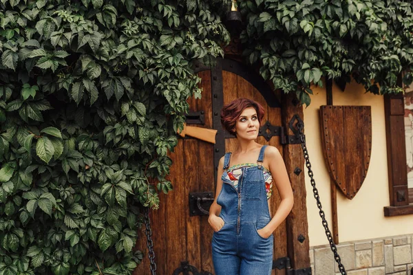 Portrait of a beautiful woman wearing denim jumpsuit standing against an ivy framed ancient wooden door of a castle. Magical toned — Stock Photo, Image