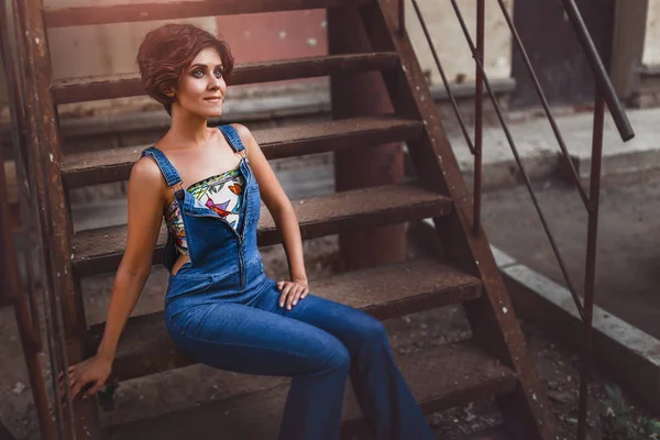 都市の若い女性トーン残忍なポーズ、珍しいブルー ジーンズの鉄階段の上に座るショット — ストック写真