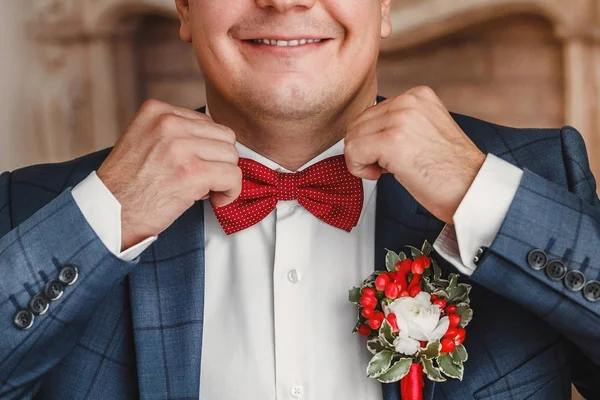 Bonito noivo endireita seu laço vermelho com boutonniere e sorrindo — Fotografia de Stock