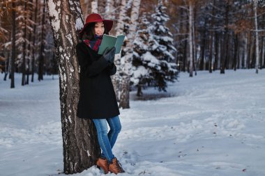 winter street portrait of young beautiful looking woman in classic clothes holding and reading book and smiling. The concept of deep and meaningful soul of a poet clipart