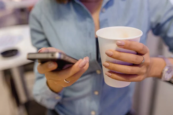 The hand of a woman with a smartphone and a takeaway mug of tea in the cafe — Stock Photo, Image