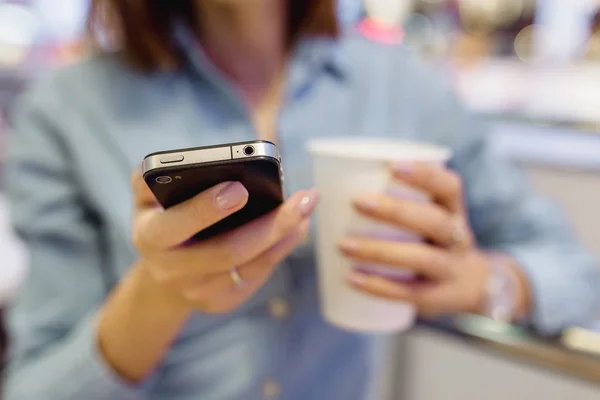 The hand of a woman with a smartphone and a takeaway mug of tea in the cafe — Stock Photo, Image