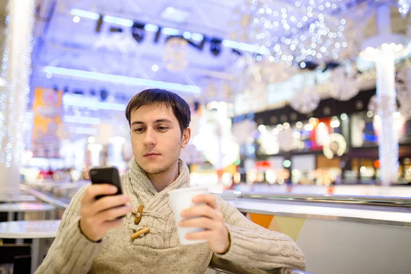 Όμορφος νεαρός άνδρας σε λευκό πουλόβερ χρησιμοποιώντας ένα έξυπνο τηλέφωνο, πίνοντας καφέ και χαμογελώντας ενώ ξεκουραζόταν σε καφέ μετά από να κάνει ψώνια στο εμπορικό κέντρο. Χριστούγεννα του χρόνου — Φωτογραφία Αρχείου