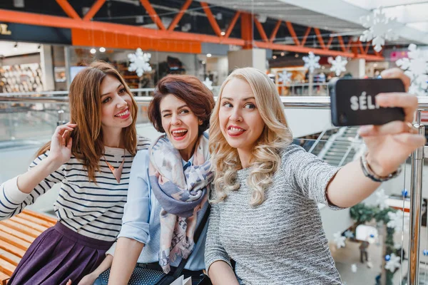 Три девушки делают перерыв в новогоднем шопинге в торговом центре и делают селфи на смартфоне — стоковое фото