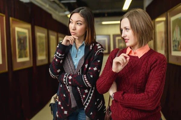 Two girl friends standing in a gallery contemplating and discuss paintings artworks displayed on gallery walls — Stock Photo, Image