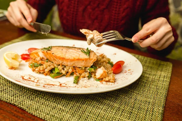 Girl with fork and knife in the hands eating salmon steak with rice and vegetables in restaurant — Stock Photo, Image