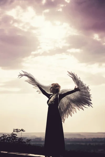 Joven mujer glamorosa en un vestido negro con un gran ángel blanco alas en el fondo de un cielo atardecer dramático. El concepto de un ángel oscuro caído — Foto de Stock