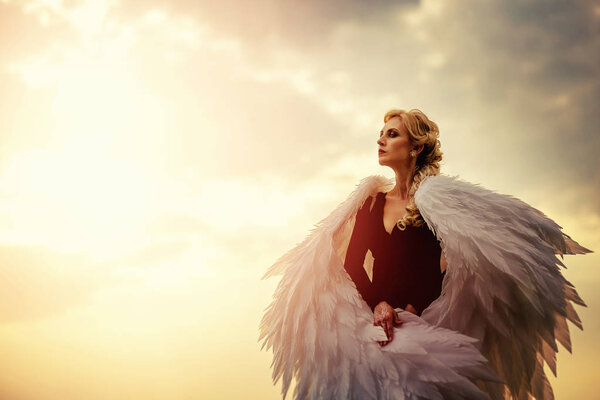 Young glamorous woman in a black dress with a large white angel wings on the background of a dramatic sunset sky. The concept of a fallen dark angel