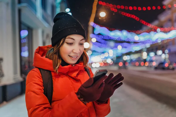 Mujer atractiva joven usando teléfono inteligente en la noche de Navidad nevada —  Fotos de Stock