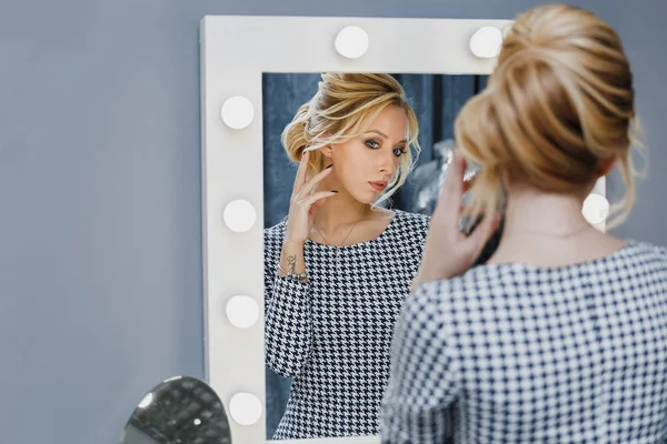 Woman in a beauty salon looks at her reflection in the mirror with lamps and checks hairstyle and makeup — Stock Photo, Image