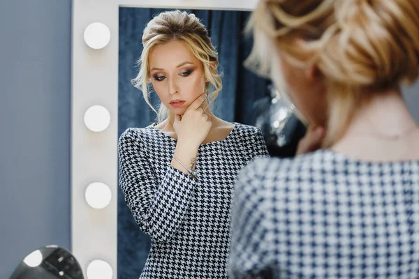 Woman in a beauty salon looks at her reflection in the mirror with lamps and checks hairstyle and makeup — Stock Photo, Image