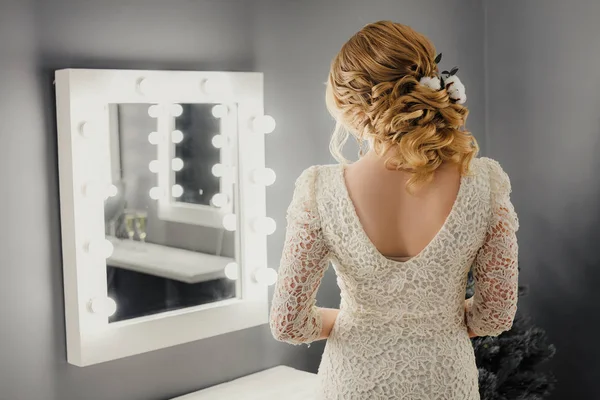 Молода красива піщано-волохата жінка зі стильною зачіскою з бавовняними квітами в бежевій сукні простежує біля дзеркала з лампами в салоні краси — стокове фото