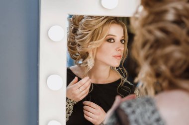 Genç güzel kadının akşam makyaj ve Güzellik Salonu lambalar ile aynaya bakarak takı ile yaratıcı saç modeli