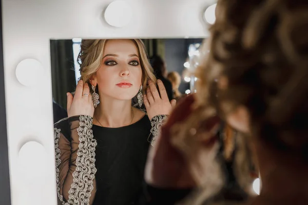 Penteado criativo de jovem mulher bonita com maquiagem à noite e jóias olhando para o espelho com lâmpadas no salão de beleza — Fotografia de Stock