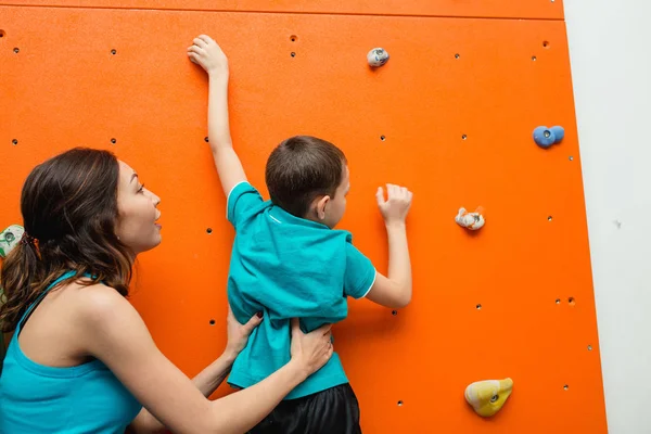 Mor att hjälpa hennes pojke att klättra på en vägg i bouldering gym på lekplats — Stockfoto