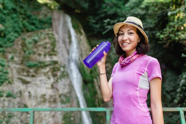 Friska hiker flicka dricksvatten i flaska framför vattenfallet. Begreppet törst — Stockfoto