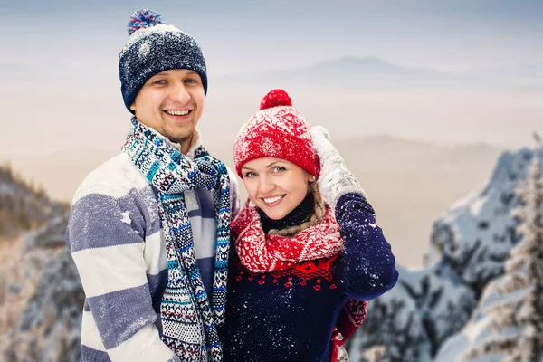 Uomo e donna sorridenti in cappelli e sciarpa e maglioni che si abbracciano sullo sfondo delle montagne invernali — Foto Stock