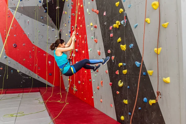 Mulher desportiva feliz com equipamento de escalada pendurado em uma corda na parede artificial de escalada interior — Fotografia de Stock