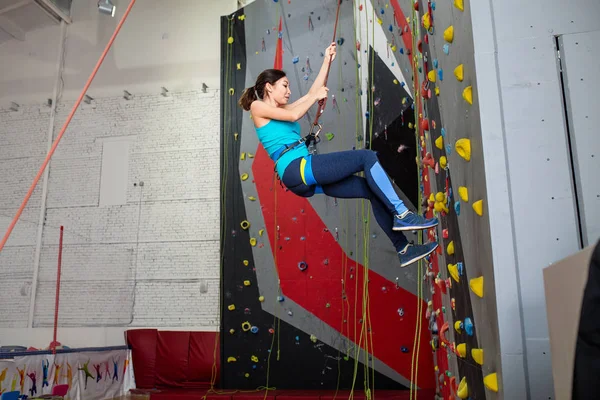 Счастливая спортсменка с альпинистским снаряжением висит на веревке у крытой скалолазания искусственной стены — стоковое фото