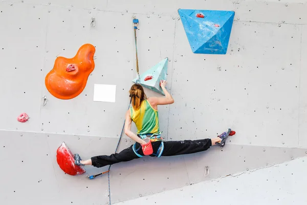 Una atleta sube la pared en un muro de escalada artificial y en un intento de tomar una ruta difícil hacer las divisiones —  Fotos de Stock
