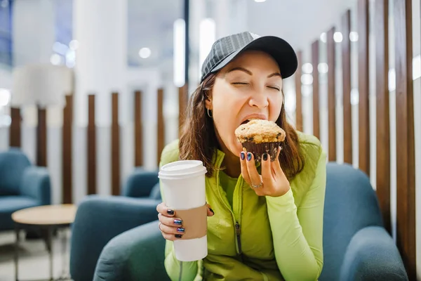 Jeune femme brune buvant du café et mangeant un délicieux gâteau au muffin dans un café d'aéroport moderne, concept de voyage fastfood — Photo