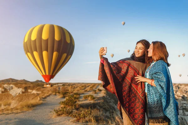 Ein paar befreundete Reisende genießen die Aussicht auf das Tal und machen ein Selfie am Telefon mit wunderbaren Ballons, die über das Tal von Kappadokien in der Türkei fliegen — Stockfoto