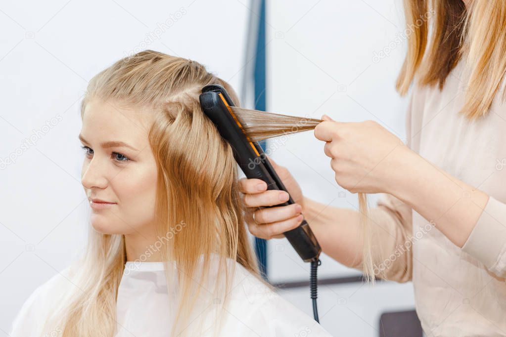 Straightening and curling of blonde hair in hairstyle salon with iron