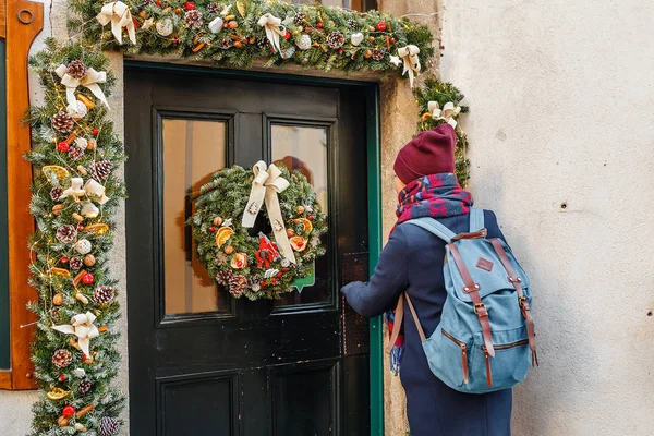 Женщина-туристка с рюкзаком, стучащая в красиво украшенную новогоднюю дверь рождественским венком — стоковое фото