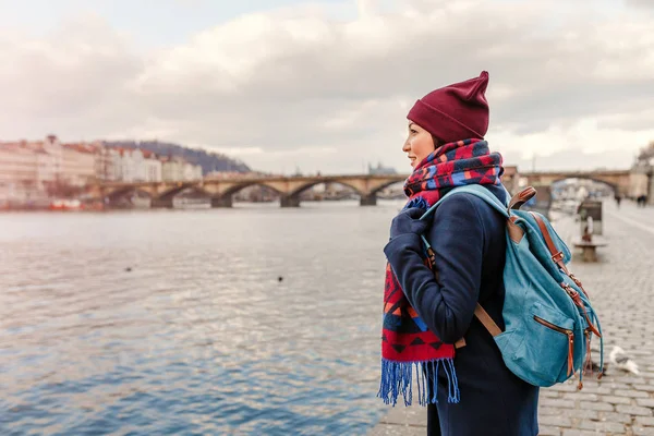 Mladá žena turista v kabátě s batoh stojící u mostu přes řeku Vltavu v Praze — Stock fotografie