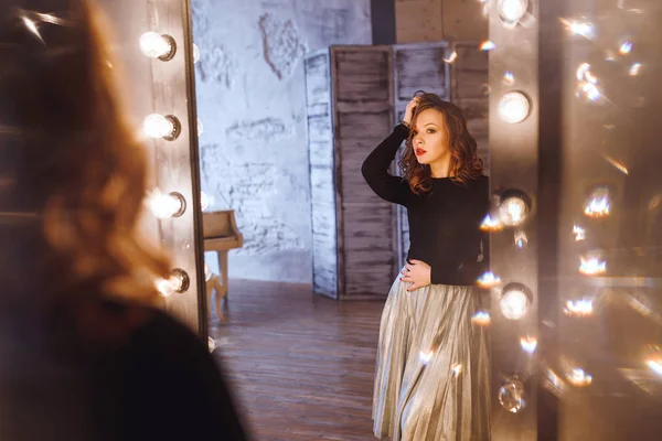 Mujer en un salón de belleza mira su reflejo en el espejo con lámparas y comprueba el peinado y el maquillaje — Foto de Stock