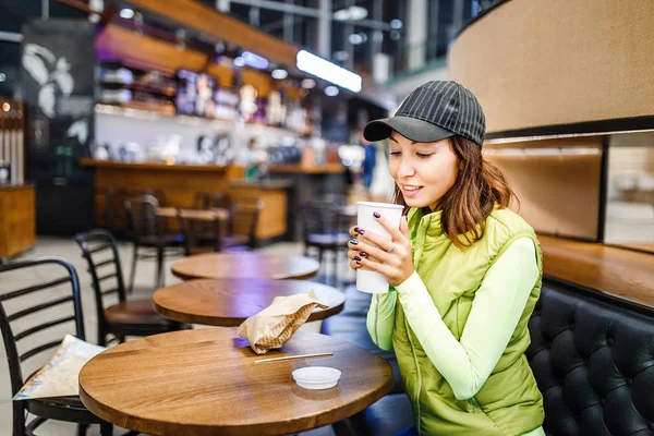 Молодая женщина пьет кофе из одноразовой чашки в кафе — стоковое фото