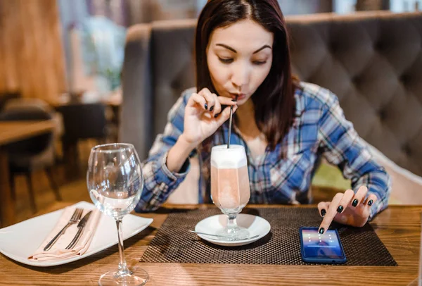 Žena sedí u dřevěného stolu v kavárně, pití kávy a držení smartphone. — Stock fotografie