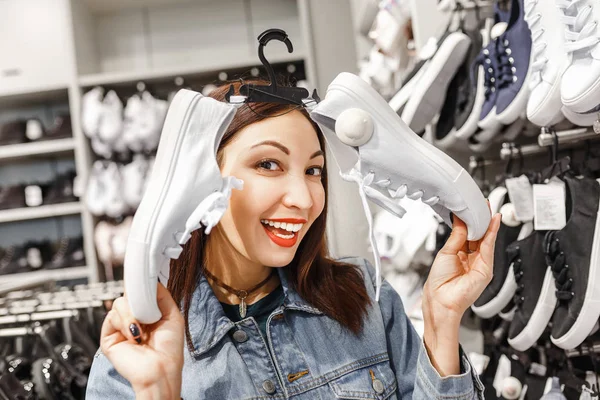 happy young woman customer choosing pair of sneakers shoes and buying it in clothes store, casual shopping concept