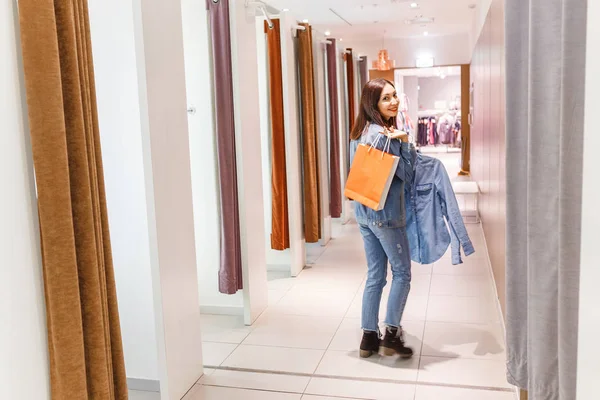 Счастливая молодая женщина примеряет одежду в гримерке и смотрит в зеркало во время шопинга в магазине моды — стоковое фото